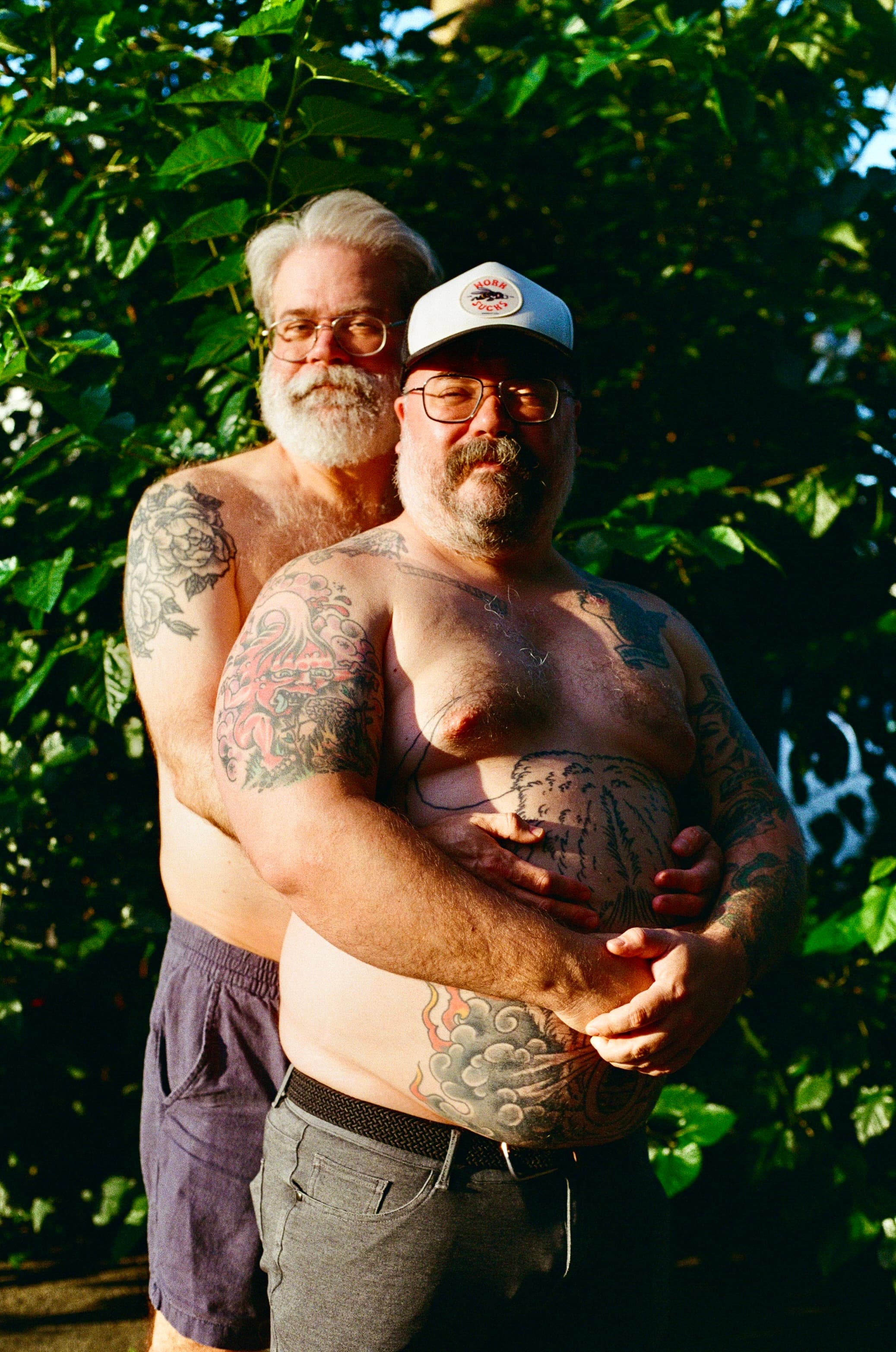 Two bearded men embracing in a sunlit garden, one resting against the other's back, both relaxed and smiling faintly.
