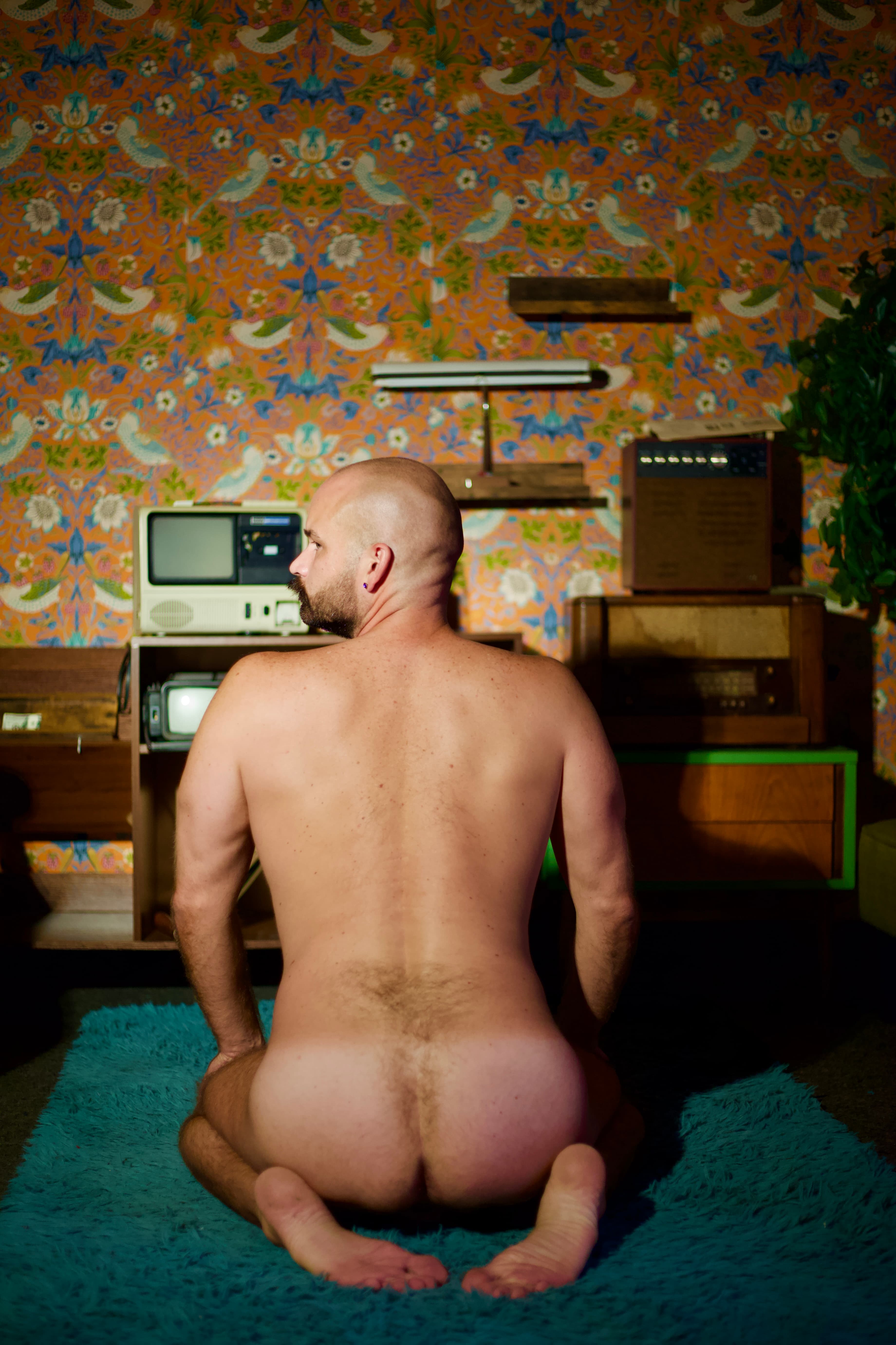 Rear view of a nude man kneeling on a teal rug in a retro room with patterned wallpaper and vintage televisions.