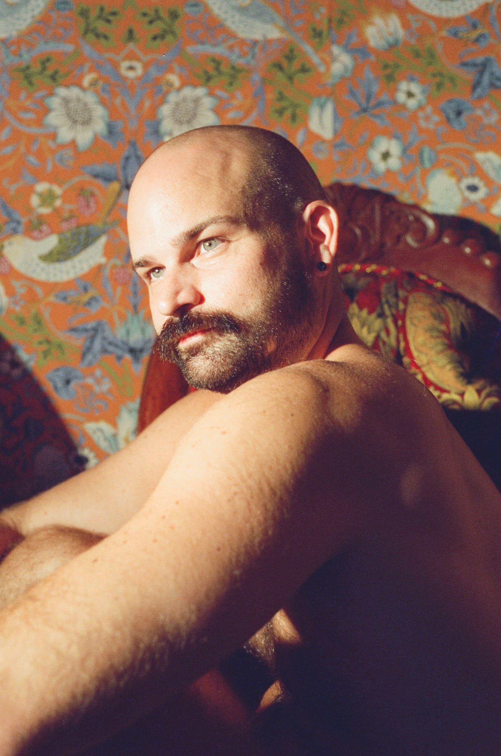 Bald bearded man reclining against ornate floral upholstery, gazing into the lens in warm afternoon light.