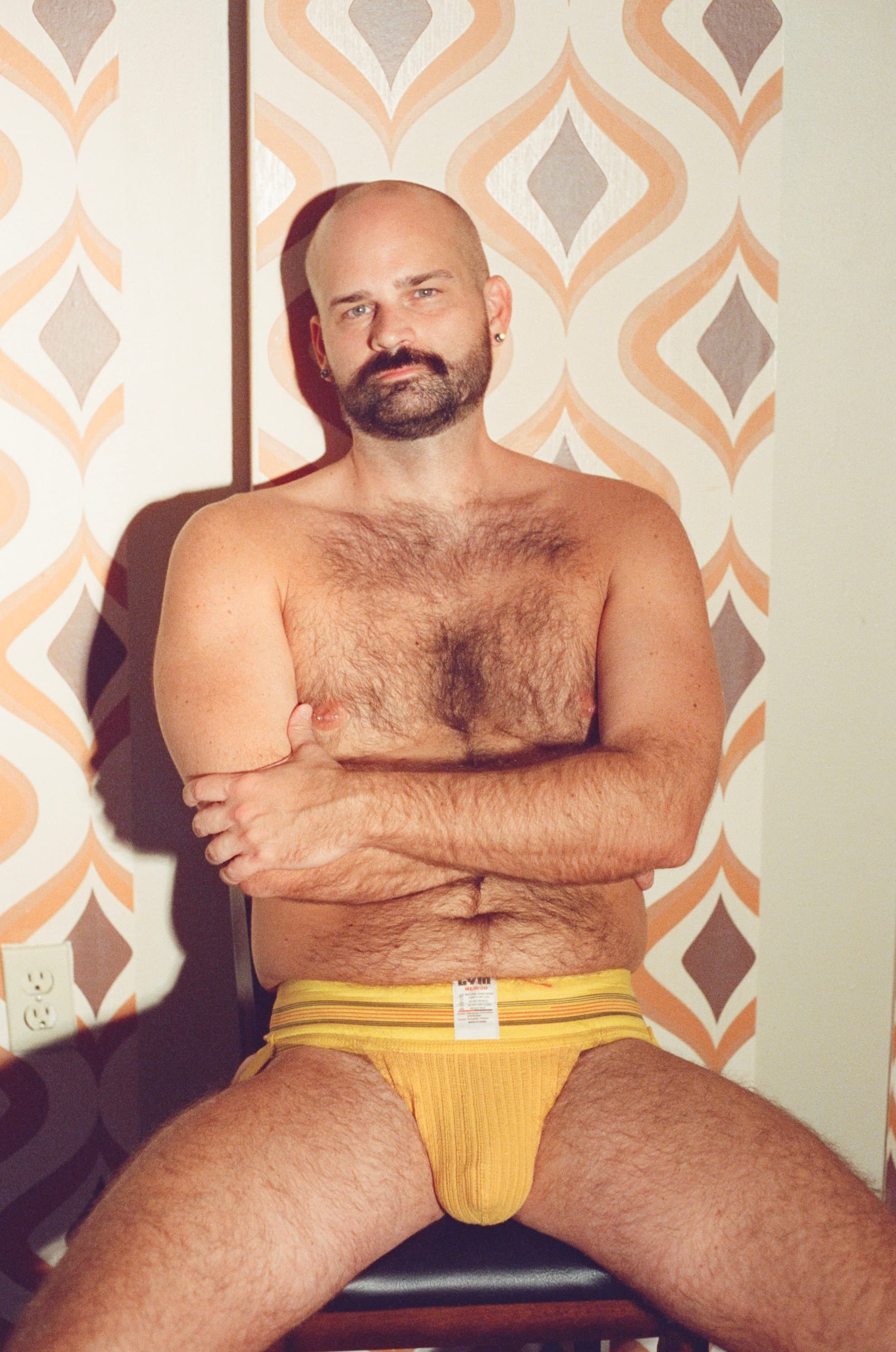 Bald bearded man seated against patterned retro wallpaper, arms crossed, steady gaze.