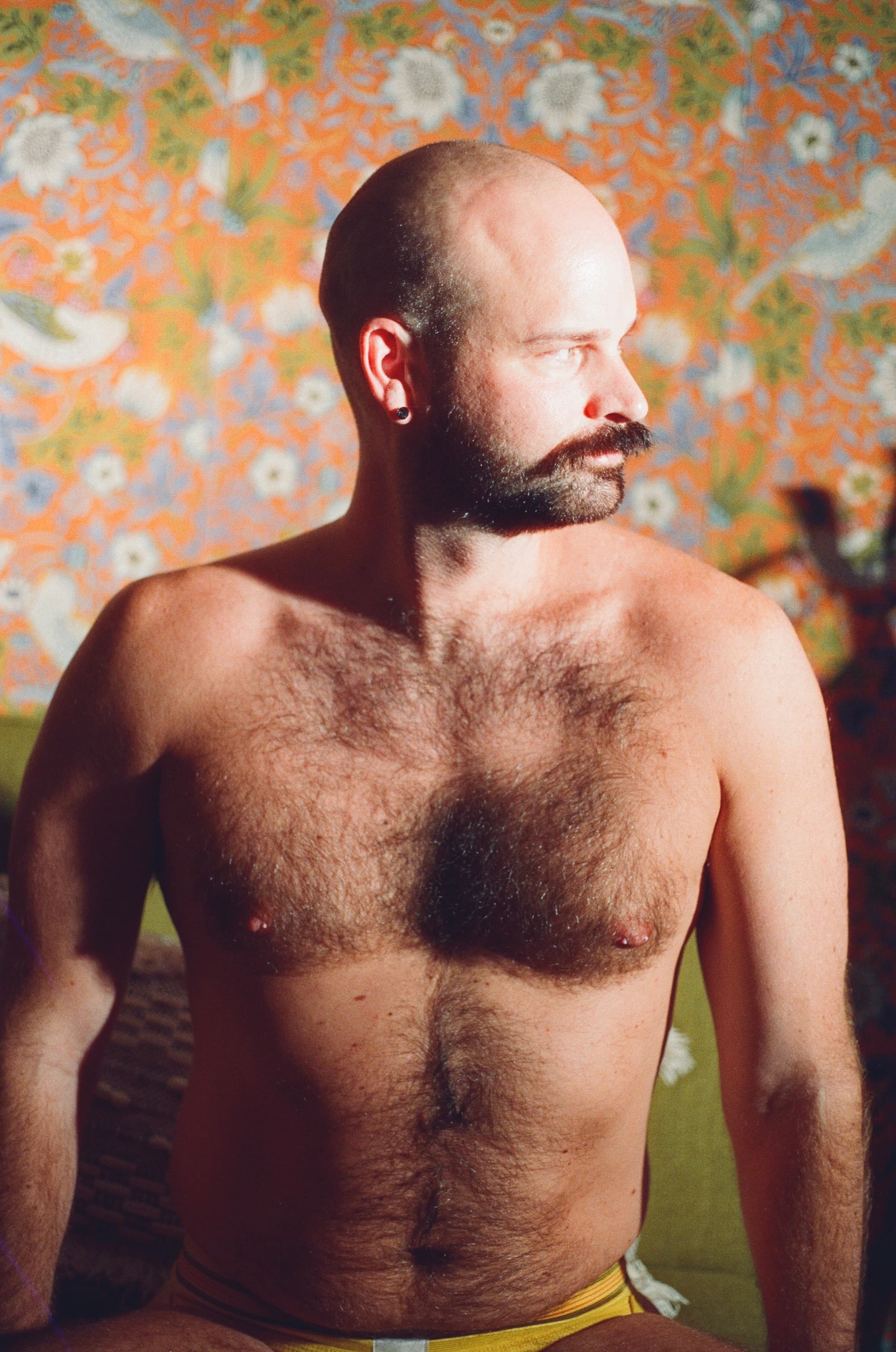 Profile of a bearded man against vivid floral wallpaper, warm directional light across the chest.