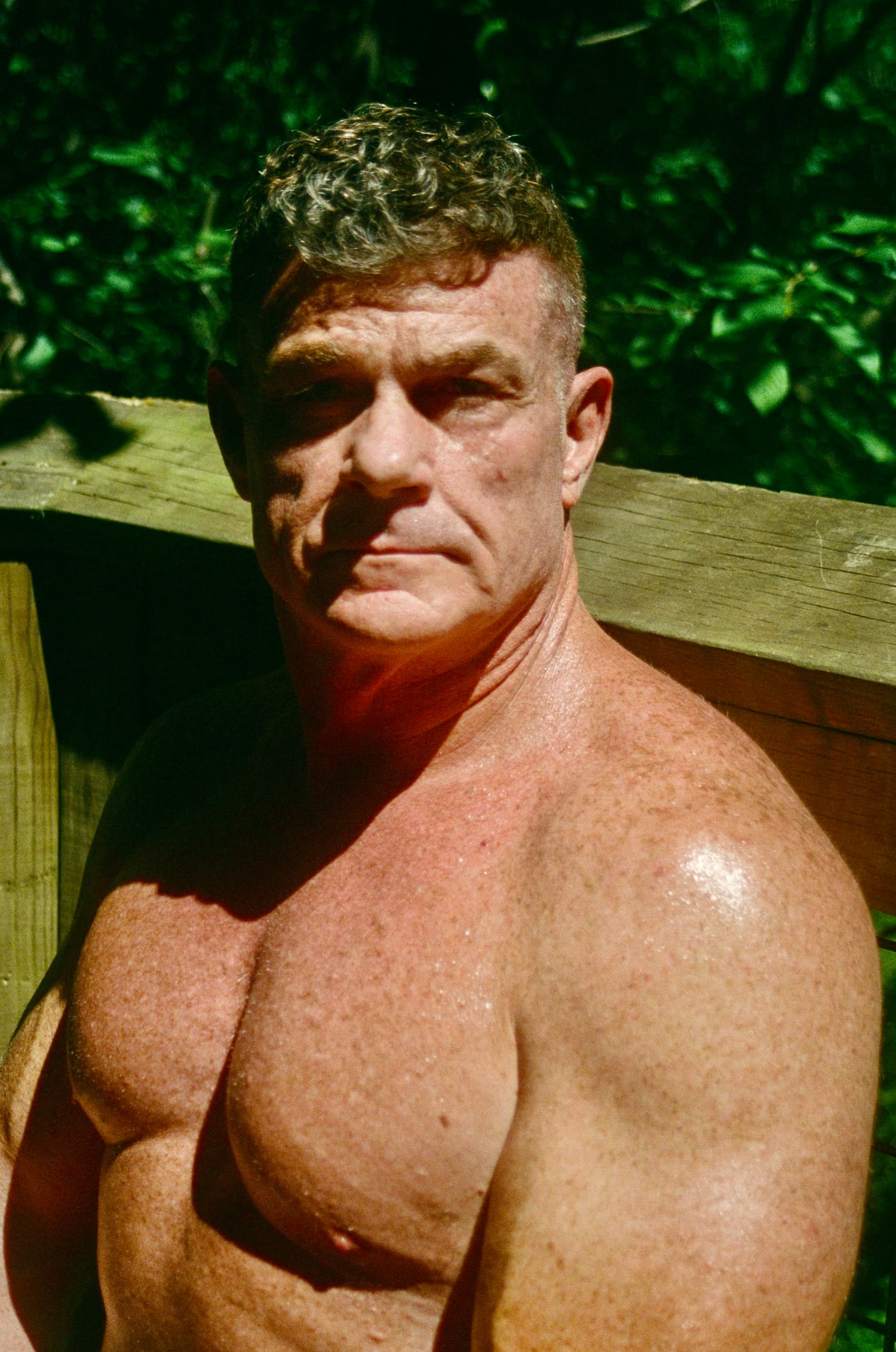 Sunlit portrait of a muscular older man with curly hair against a wooden fence and greenery.