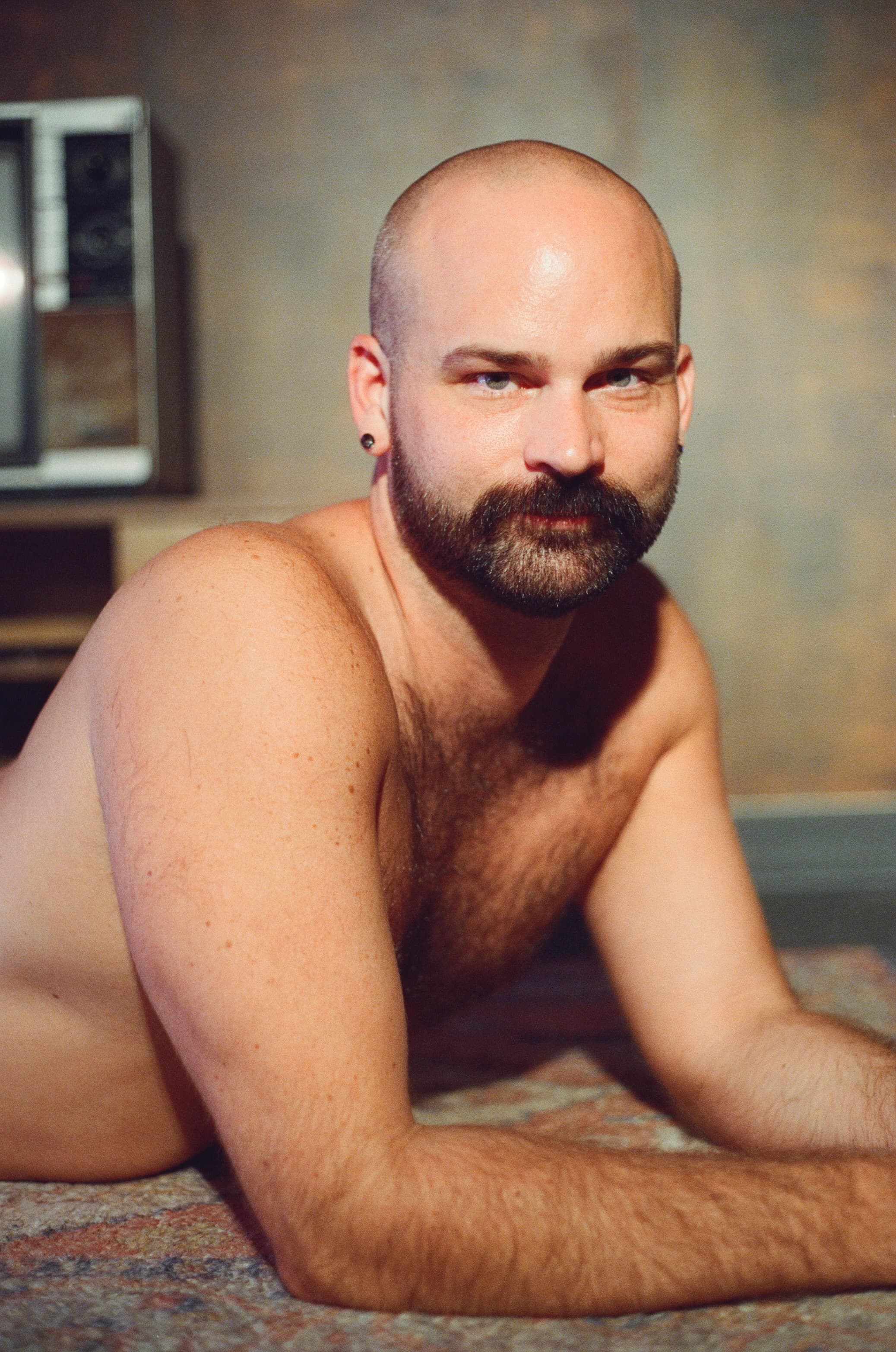 Bald bearded man reclining on a patterned rug, relaxed and looking to camera in warm light.