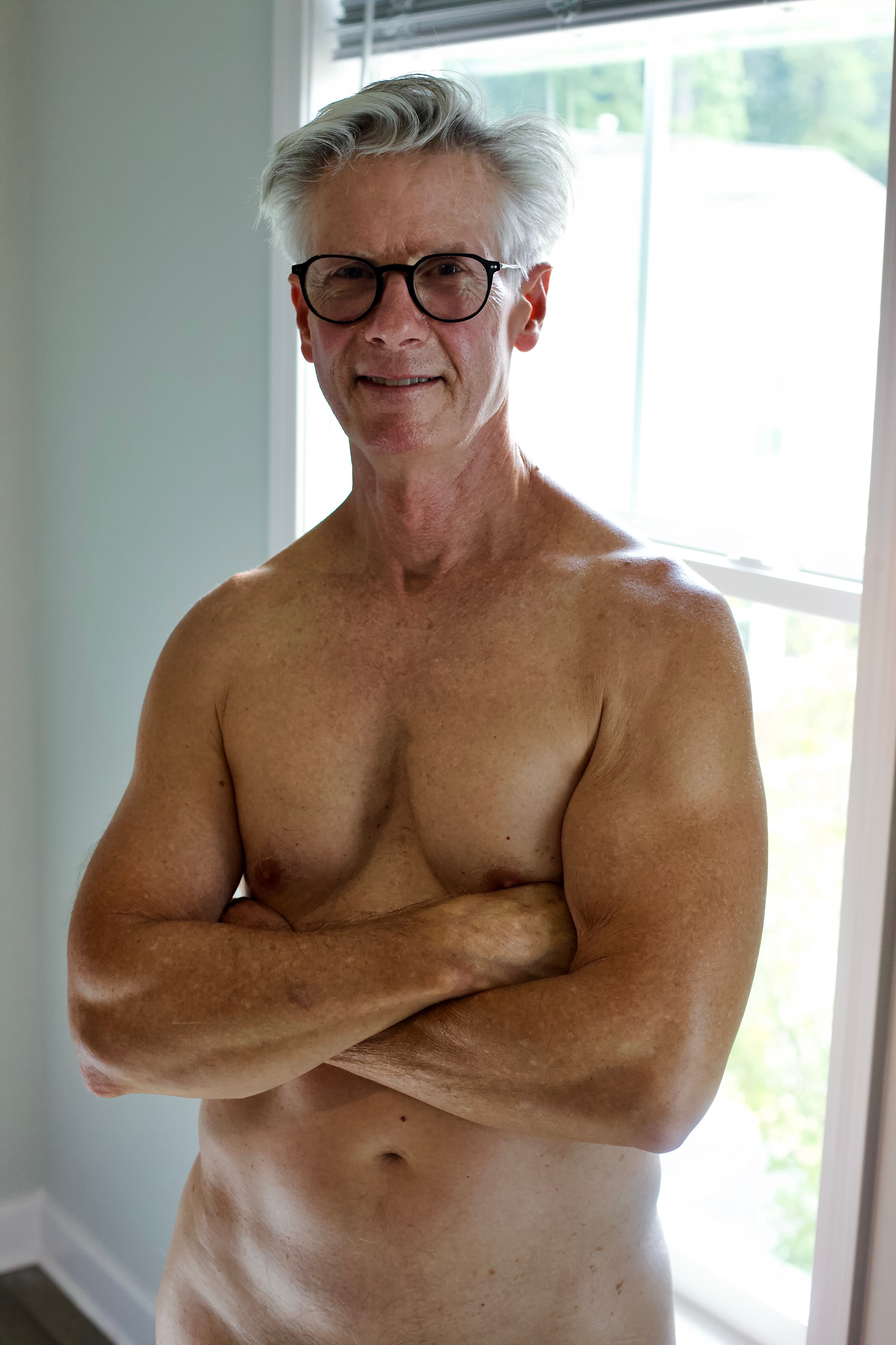 Silver-haired man in glasses standing with arms crossed by a bright window, relaxed smile in soft daylight.